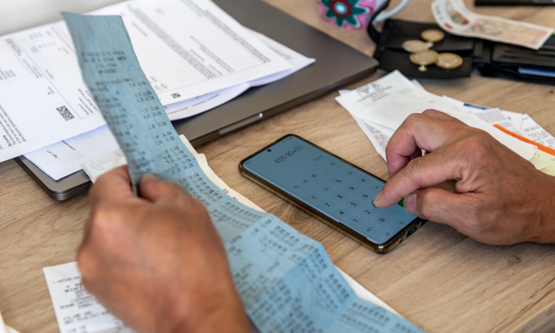 A person at a desk, adding amounts from bills and receipts. There are some coins and notes on the desk.