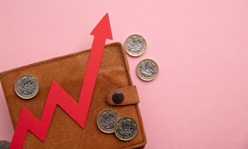 A wallet with a few pound coins around, on a pink background, with an arrow going up indicating a rise in price.