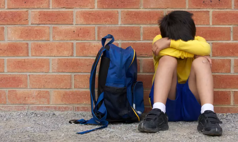 A young boy sitting knees up against a wall, his head hidden in his arms.