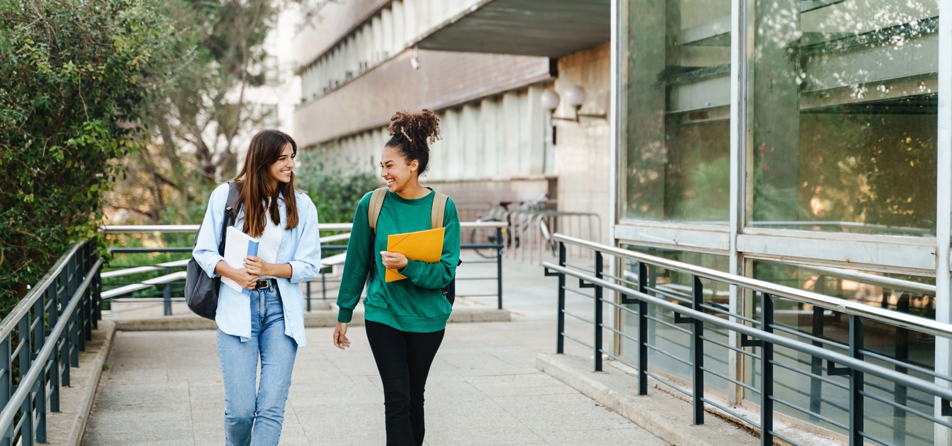 Two teen girls walking and chatting with books in their hands near a school.