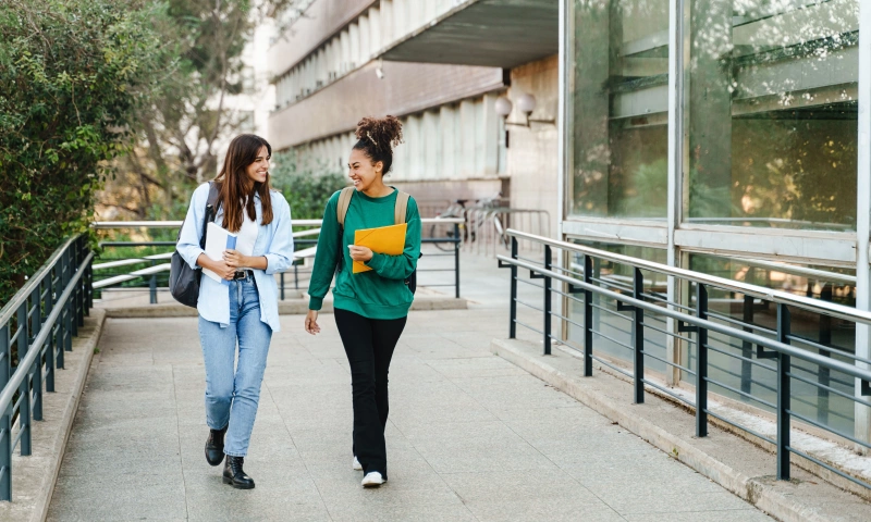 Two teen girls walking and chatting with books in their hands near a school.