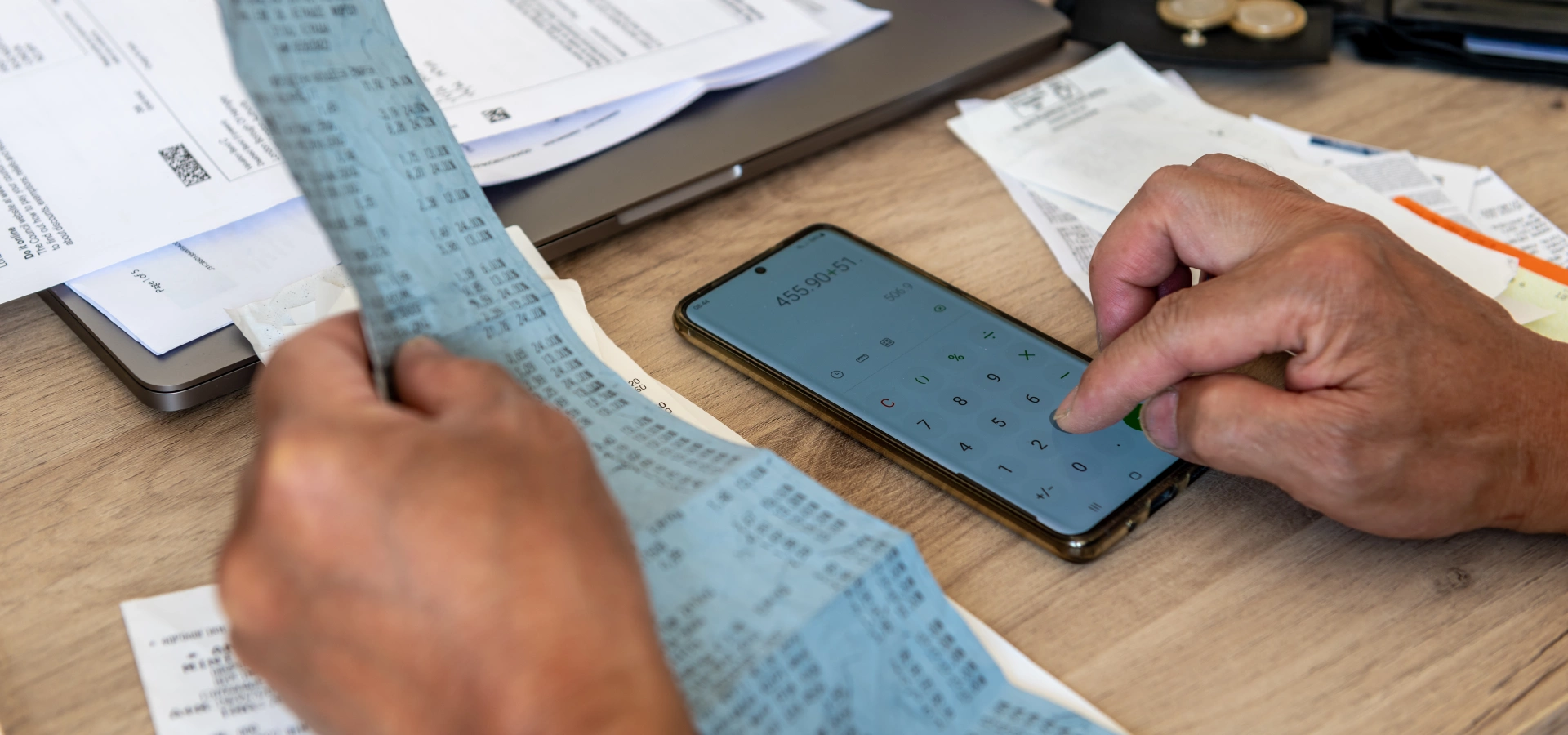 A pair of hands, one holding a very long receipt, the other tallying the total on a calculator. There are coins around.