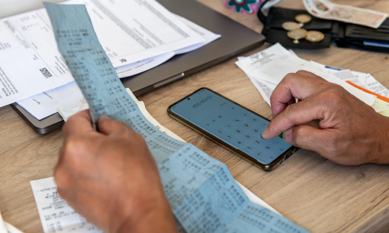 A pair of hands, one holding a very long receipt, the other tallying the total on a calculator. There are coins around.