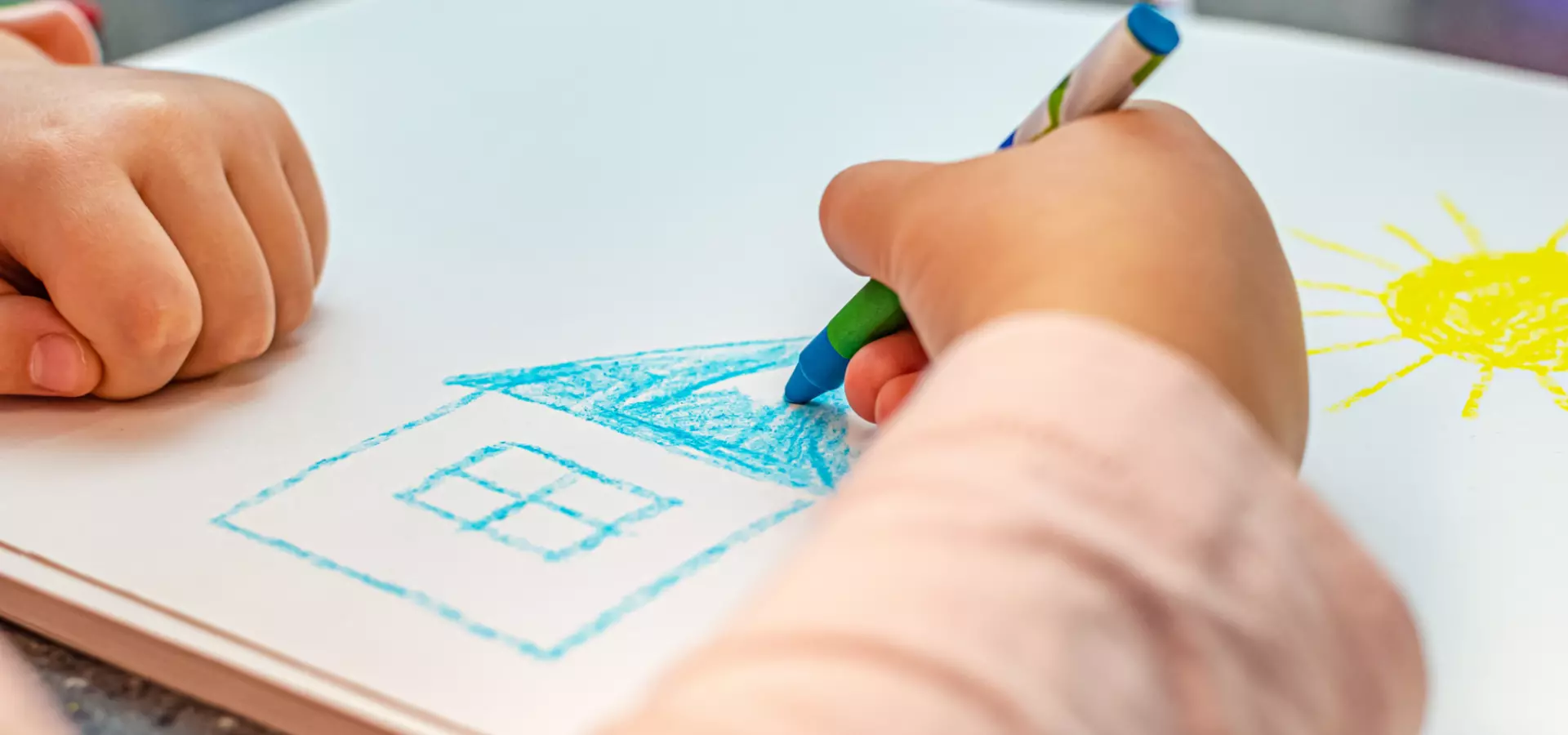 Close up of the hands of a child, drawing a house on a piece of white paper.