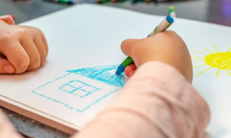 Close up of the hands of a child, drawing a house on a piece of white paper.