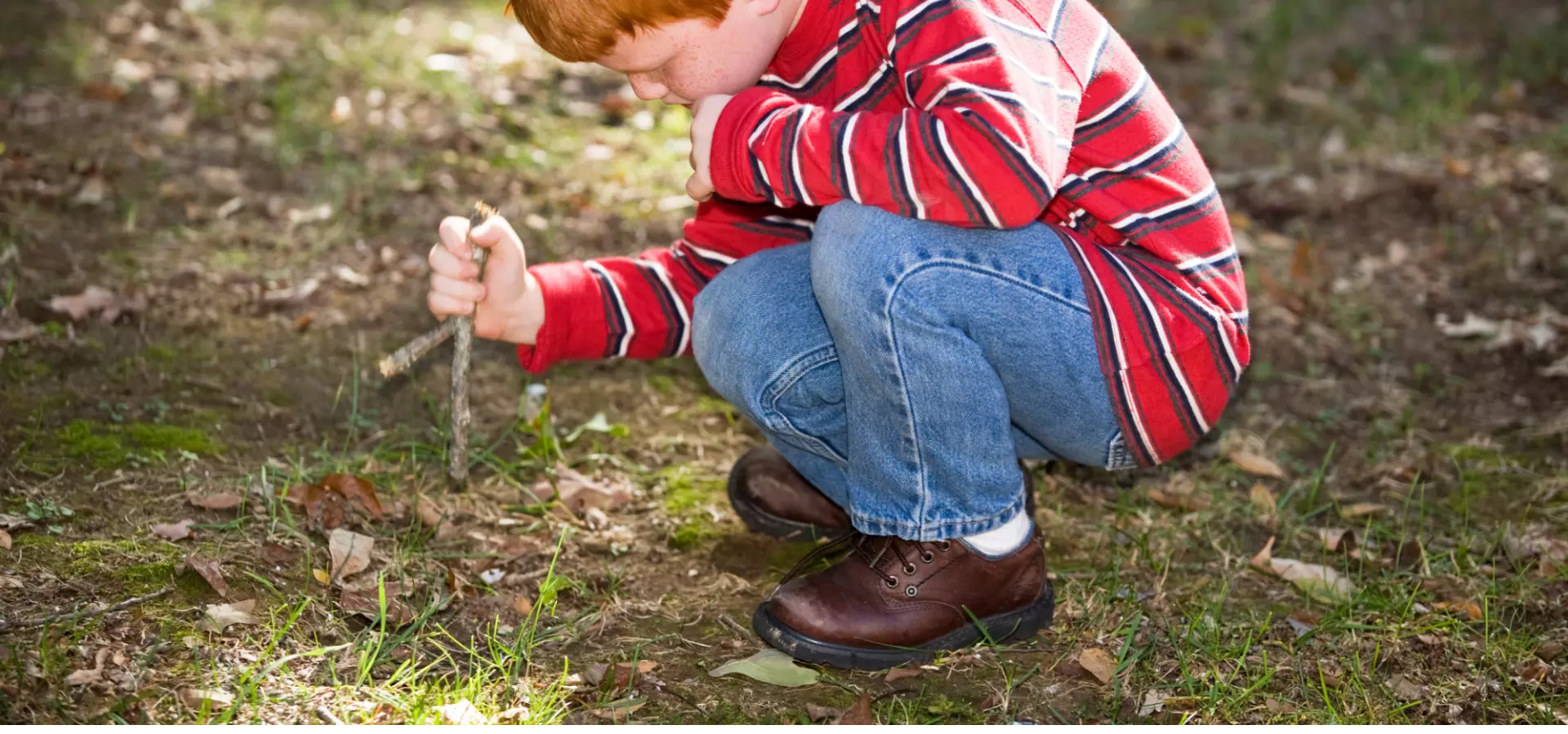 A young boy playing on his own outdoors. He is holding a stick and digging the ground with it.