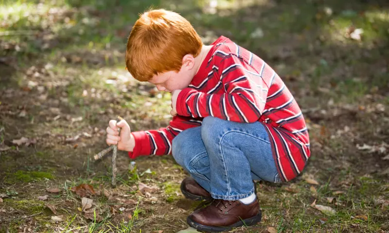 A young boy playing on his own outdoors. He is holding a stick and digging the ground with it.