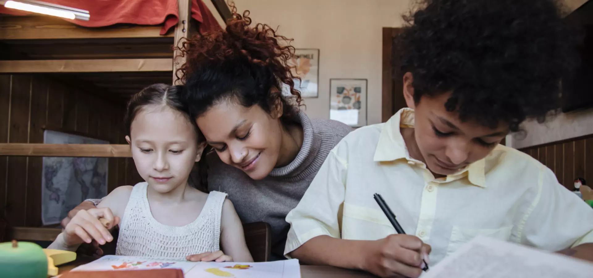 Mother sitting at a table with her two children. She is hugging her daughter, both children and reading or doing their homework.