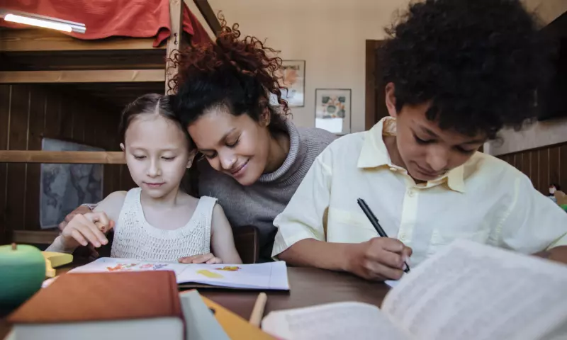 Mother sitting at a table with her two children. She is hugging her daughter, both children and reading or doing their homework.