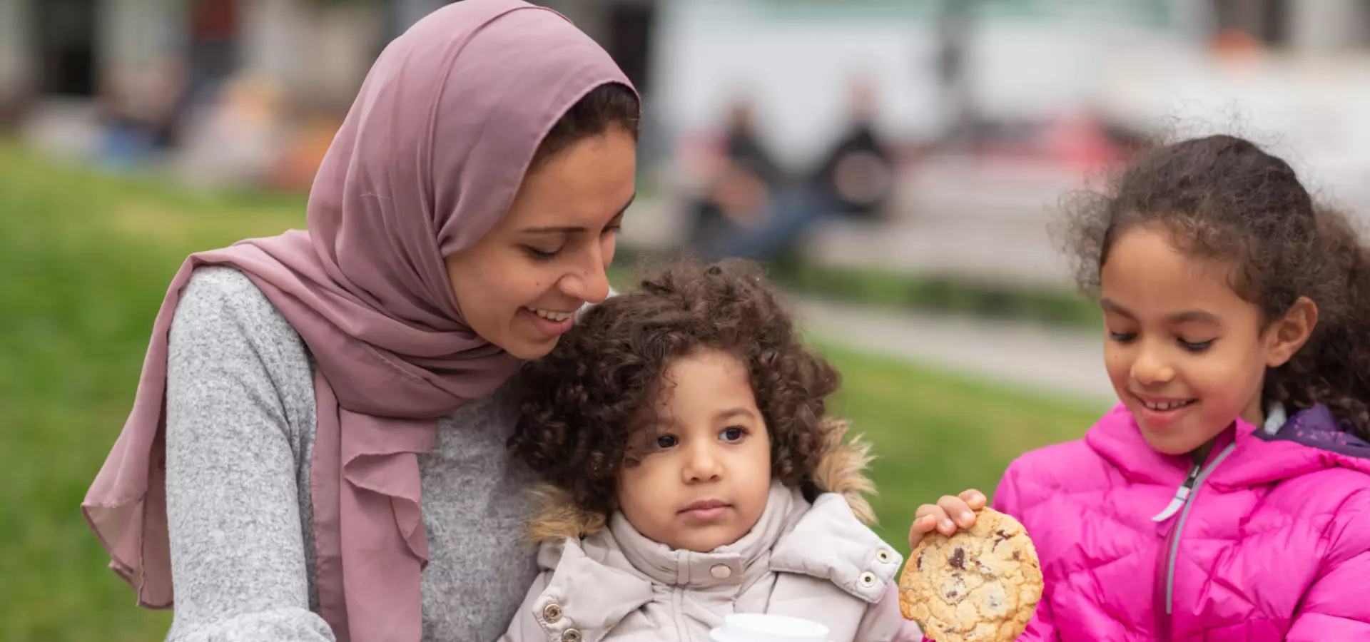 A woman wearing a head scarf sitting on a bench with her two children. She is drinking coffee in a take away cup and her children are eating cookies.