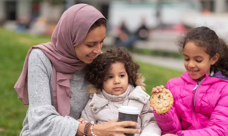 A woman wearing a head scarf sitting on a bench with her two children. She is drinking coffee in a take away cup and her children are eating cookies.