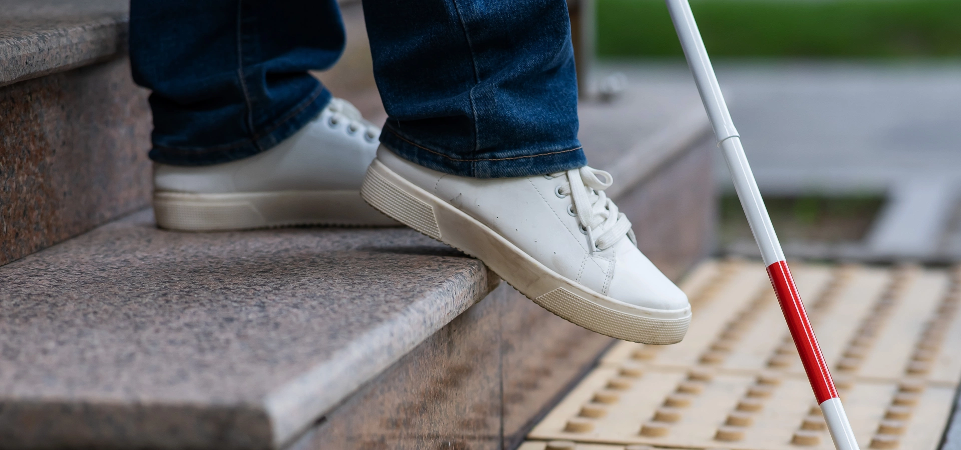 Close up on the legs and feet of a person walking with a white stick usually used by people with blindness.