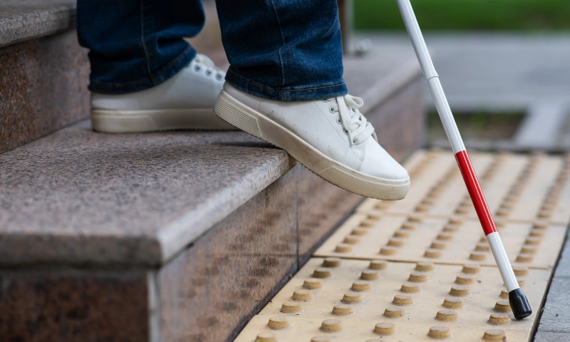 Close up on the legs and feet of a person walking with a white stick usually used by people with blindness.