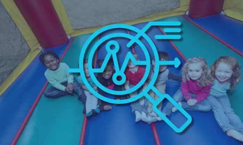 Diverse group of children sitting down on the ground and looking up at the camera, smiling. There is a magnifying glass icon on top, signifying 'overview'. 