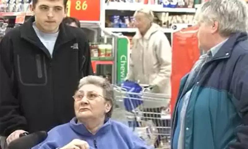 An older woman in a wheelchair is being interviewed, with a young man behind her and another person on her left. They seem to be in a supermarket.