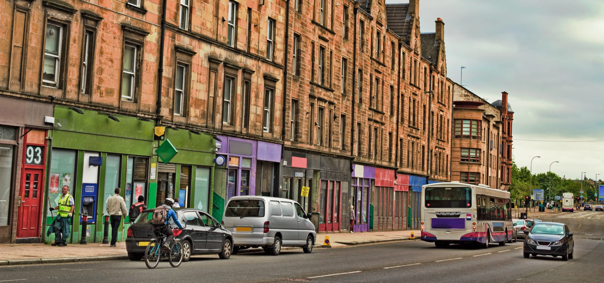 A street in Glasgow with red stone tenements, shop fronts, and parked cars.