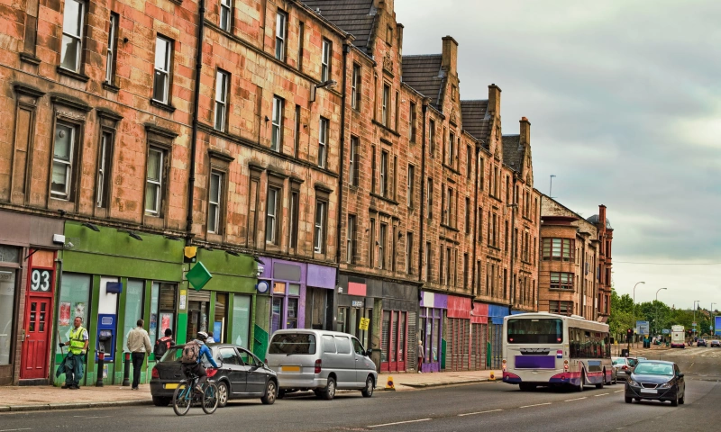 A street in Glasgow with red stone tenements, shop fronts, and parked cars.