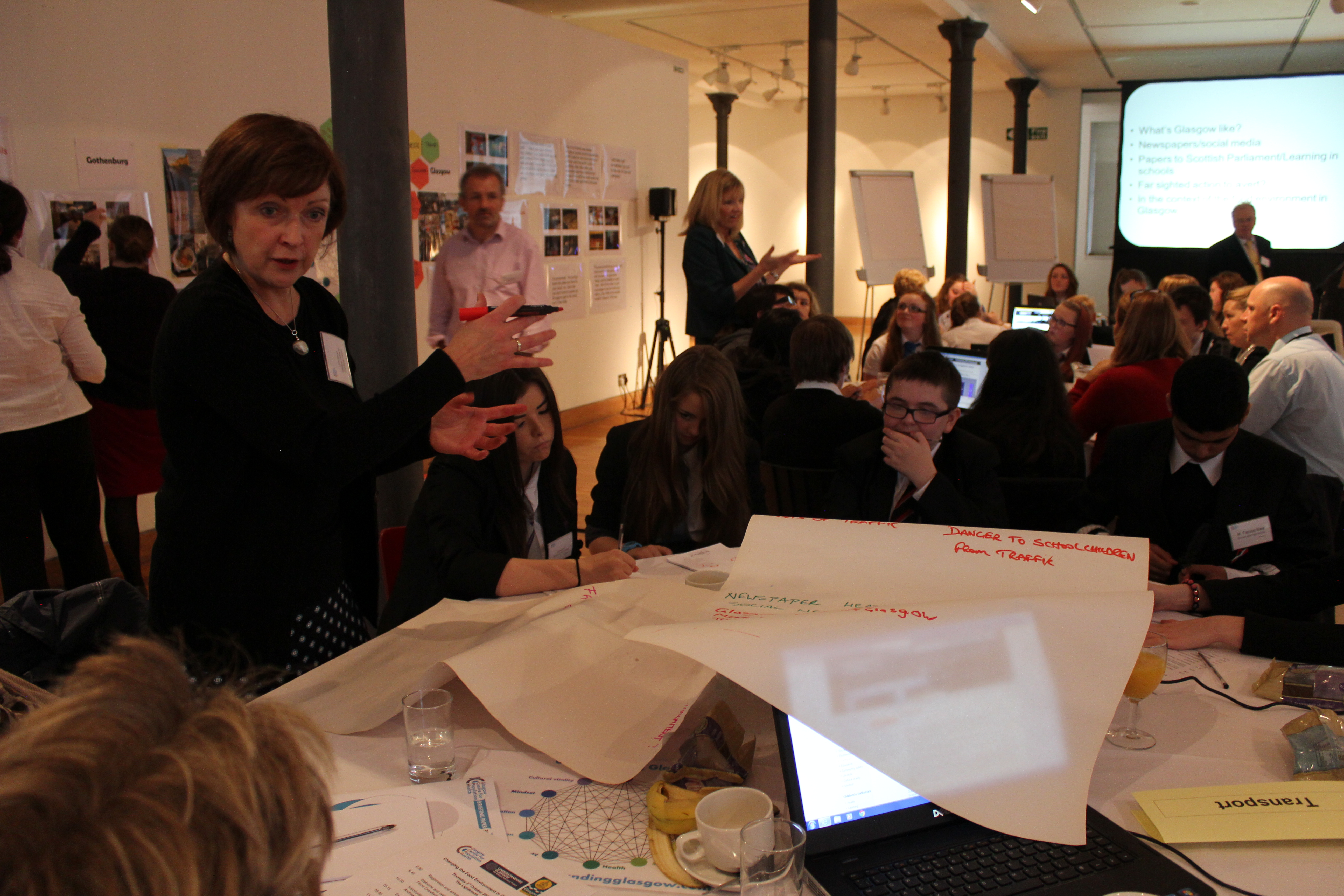 A researcher standing by a table of people, leading a discussion. Other tables in the background, with notes on tables and on the walls. 