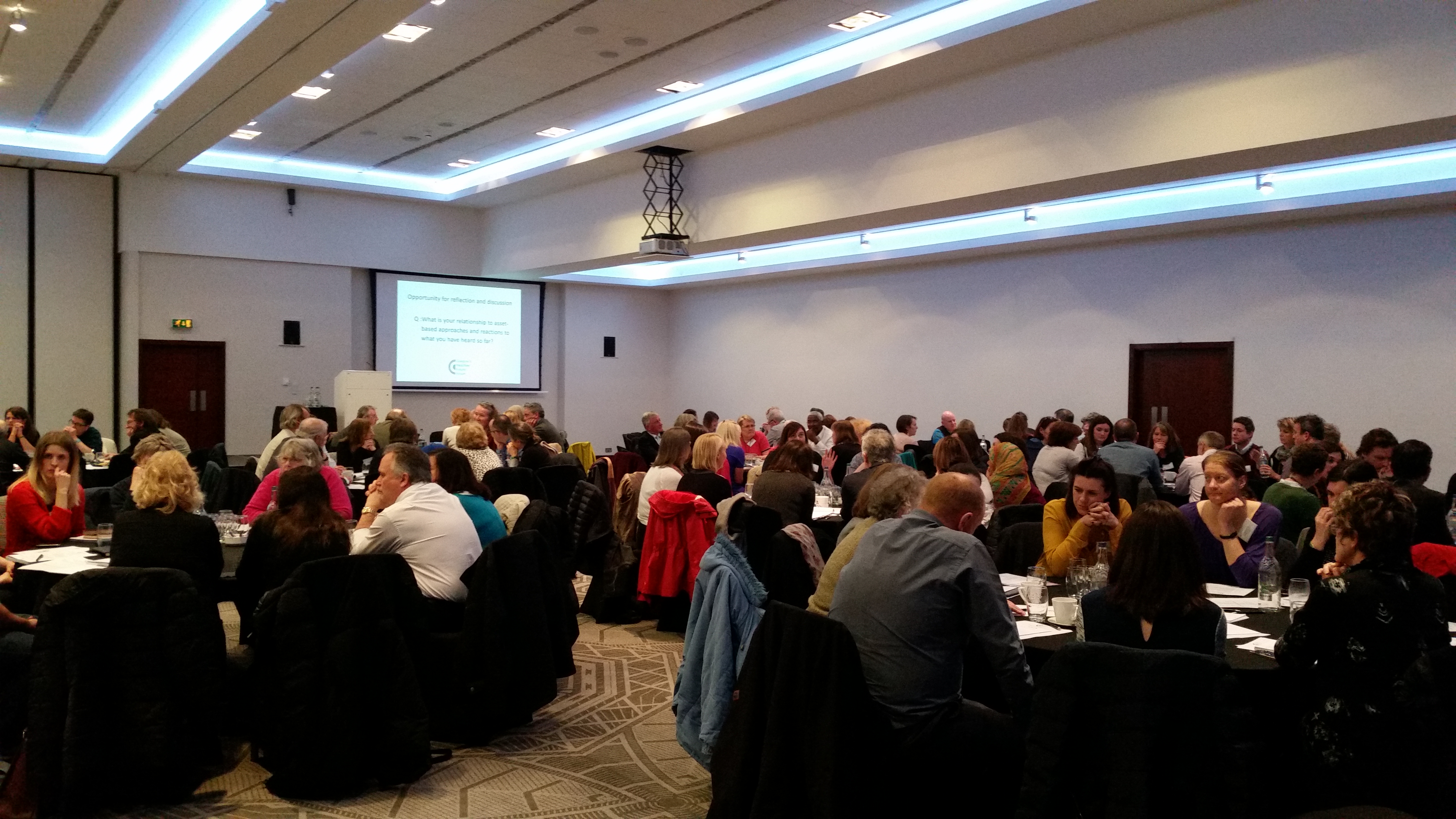 A room of people sitting at tables with a presenter at the front standing by a screen.